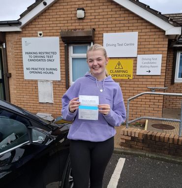 Young woman proudly holds her practical driving test pass certificate outside a test centre.