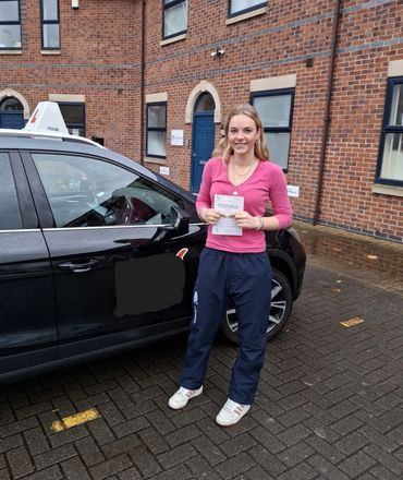 Young woman proudly holding a practical driving test pass certificate next to a learner driver car.