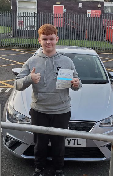 Teen proudly holds driving test pass certificate in front of silver car.