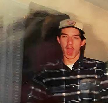 Young man with a mustache wearing a cap and striped shirt, mid-expression.