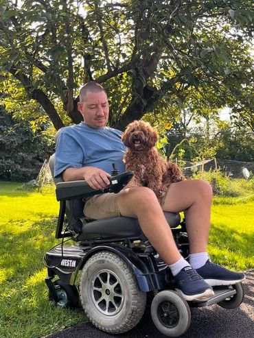 Man in a wheelchair with a happy brown dog on his lap in a sunny garden.