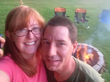 Smiling couple takes a selfie near a fire pit outdoors.