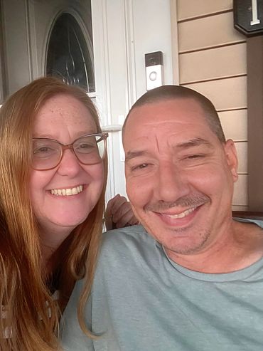 Smiling couple taking a selfie on a porch with a doorbell visible.