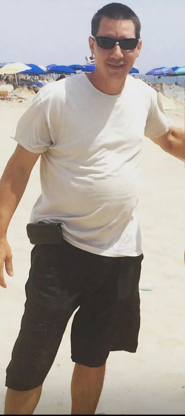 Man wearing sunglasses and casual clothes on a sunny beach with umbrellas in the background.