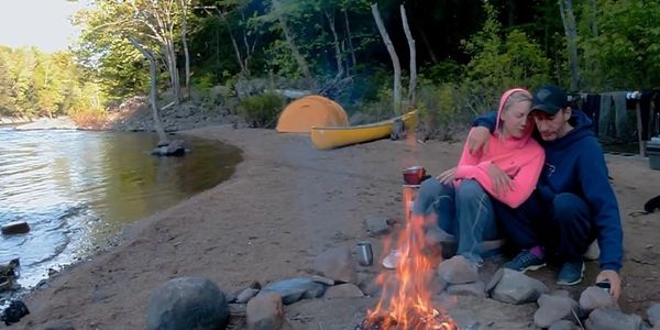 Couple cuddling by a campfire near a lakeside tent and canoe.