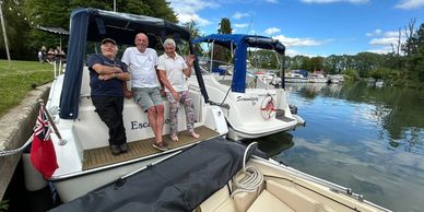 Three men relaxing on a docked boat by a calm river under a partly cloudy sky.