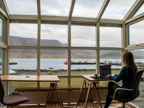 A woman working on a laptop in a room with large windows overlooking a lake and mountains.