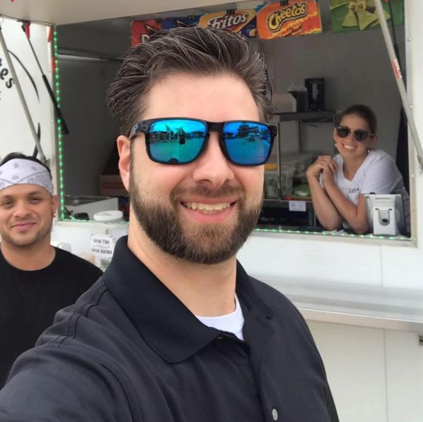 Three people smiling at a food stand, with one man in sunglasses taking a selfie.