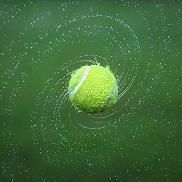 Wet yellow tennis ball with water droplets spiralling out from the centre.