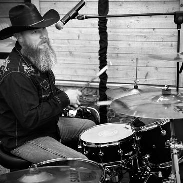 Bearded man in cowboy hat passionately playing drums.