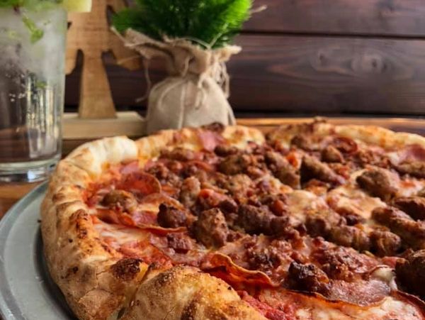 Close-up of a meat-topped pizza on a metal tray with a drink and plant in the background.