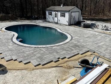 A backyard pool surrounded by newly laid stone tiles, with a shed in the background and construction tools nearby.