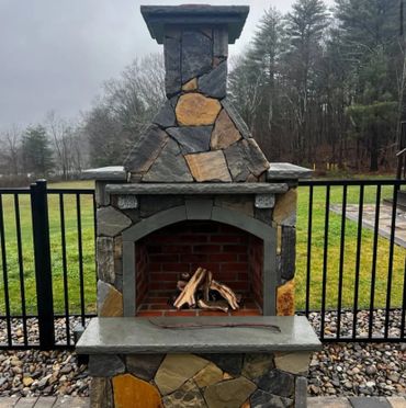 Outdoor stone fireplace with wood logs ready to ignite, set against a green yard and black fence.