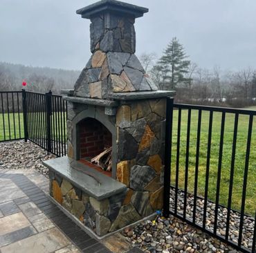 Outdoor stone fireplace on a patio with a metal fence and a grassy field in the background.