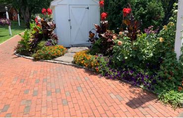 A brick pathway leads to a white shed surrounded by colorful flowers and greenery.