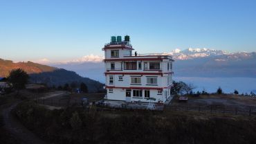 A white multi-story house with red trim stands against snowy mountain peaks under a clear blue sky.