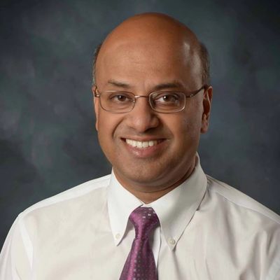Smiling man in glasses and a tie against a dark background.