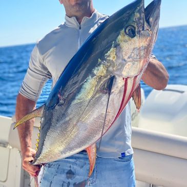 Man with sunglasses hoisting up a big yellowfin tuna in his arms on a boat.