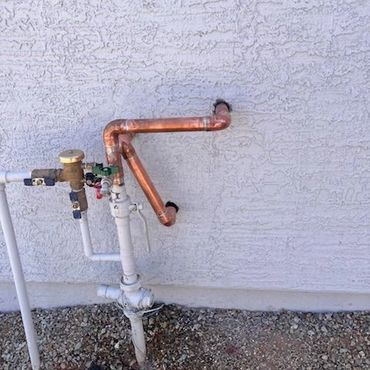 Copper and white pipes attached to a textured white wall above rocky ground.