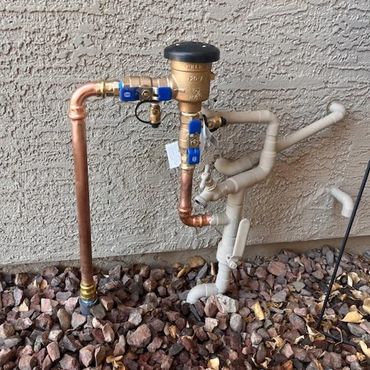 Copper and PVC pipes connected to a brass valve on a rocky ground against a textured wall.