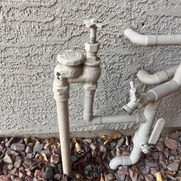 Outdoor plumbing pipes and valves against a textured wall with gravel base.