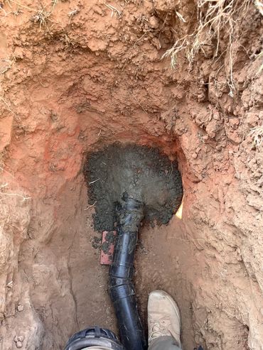 A black pipe is installed in a dirt trench with a boot nearby for scale.
