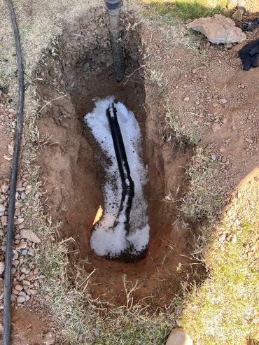 Excavated trench with black pipes and white gravel bedding for plumbing installation.