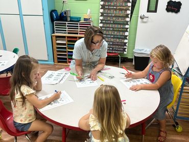Teacher helps young children with worksheets at a round table in a colorful classroom.