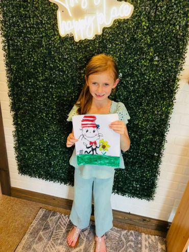 A young girl proudly holds her colorful drawing in front of a leafy green backdrop.