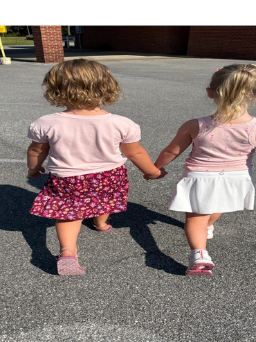 Two young girls holding hands walking on pavement outside.