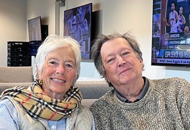 Medieval murder mysteries Authors Lady and the Doc smiling indoors with TV screens in the background.