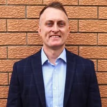 Man in a blue suit jacket and light blue shirt standing against a brick wall.