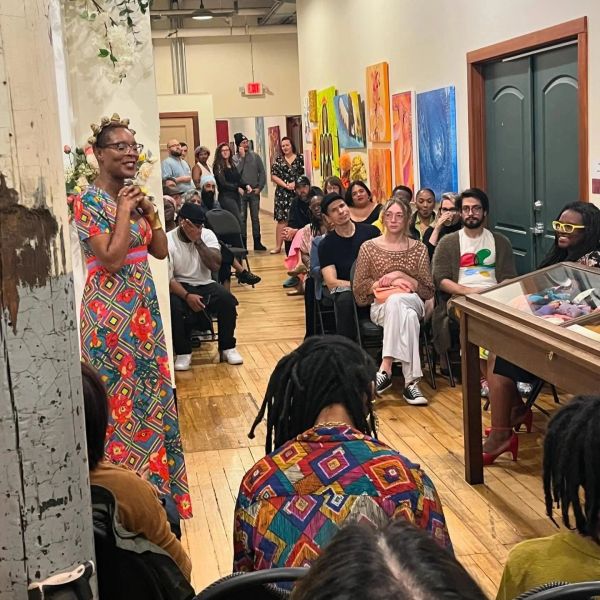 A woman in a colorful dress speaks to an attentive audience in an art gallery.