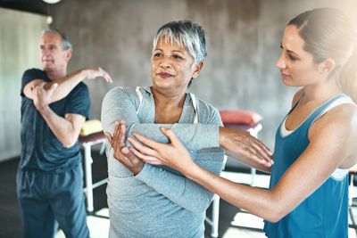 A fitness instructor assists a middle-aged woman with arm stretches in a gym setting.