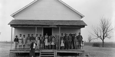 Children and a teacher stand outside an old rural schoolhouse.