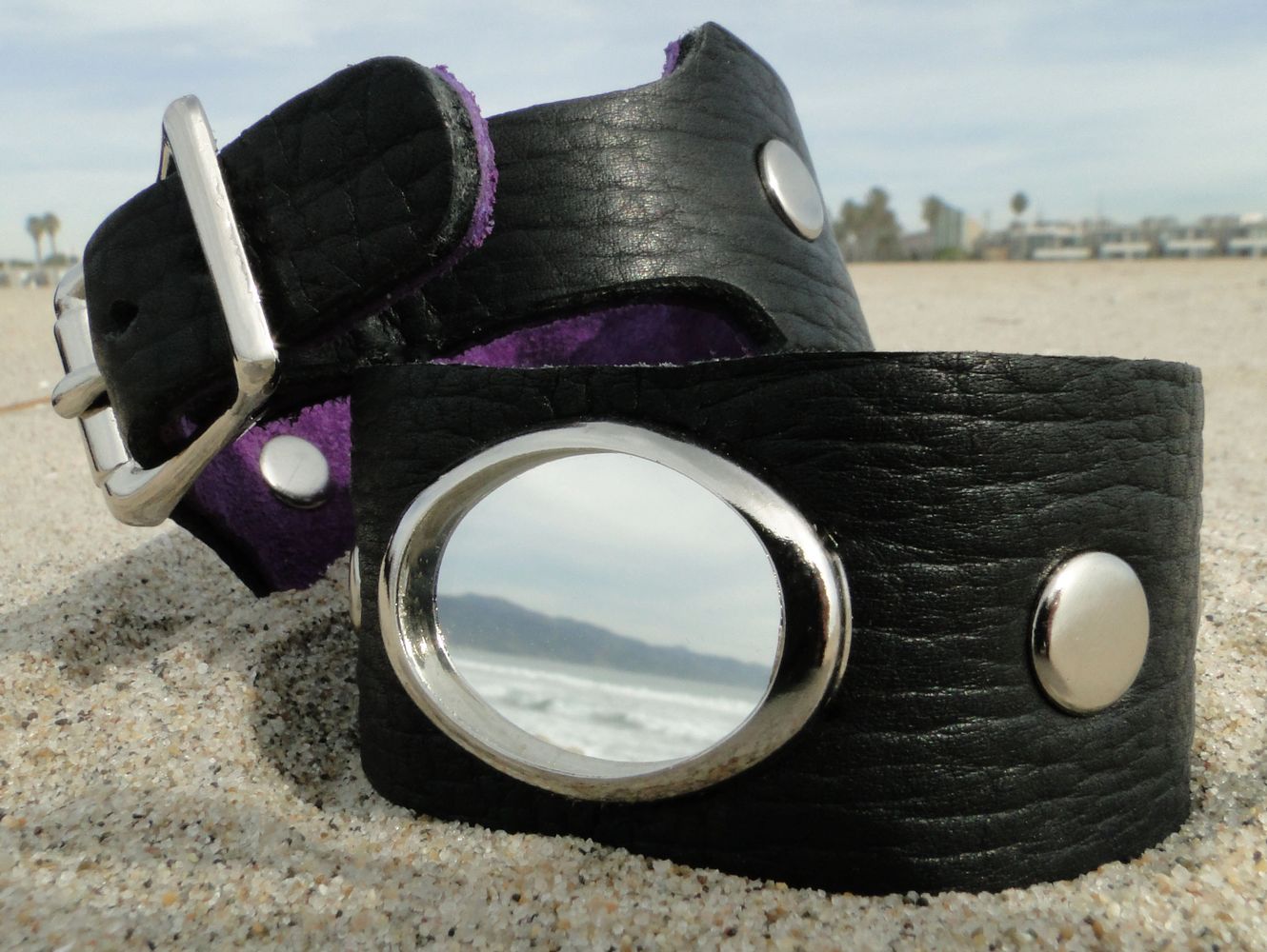 Close-up of a black leather bracelet with metal studs and a circular mirror on a sandy beach.