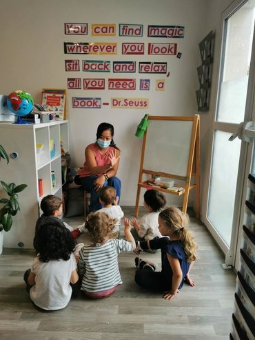 Teacher wearing a mask engaging young children in a classroom with a Dr. Seuss quote on the wall.