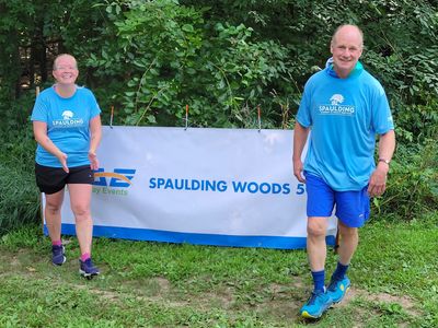 Two people in blue shirts standing near a Spaulding Woods 5K banner outdoors.