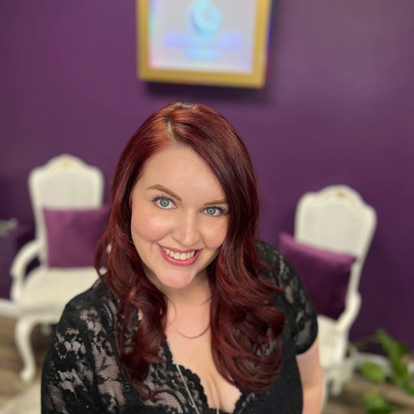 Smiling woman with red hair in a black lace top against a purple background.