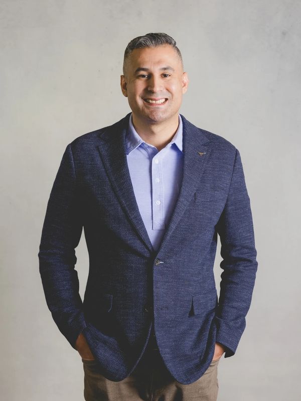 Smiling man in a blue blazer and light blue shirt stands confidently with hands in pockets.