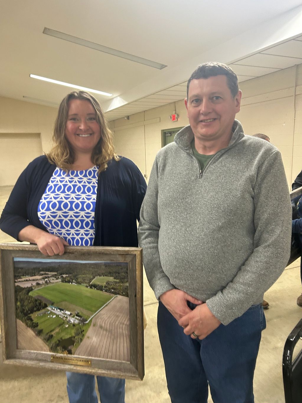 Two people smiling, one holding a framed aerial photo of farmland.