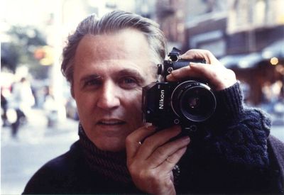 Color photograph of  Gerard Malanga holding a 35mm camera. 