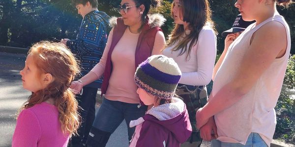 A group of people standing outdoors in sunlight, dressed warmly and casually.