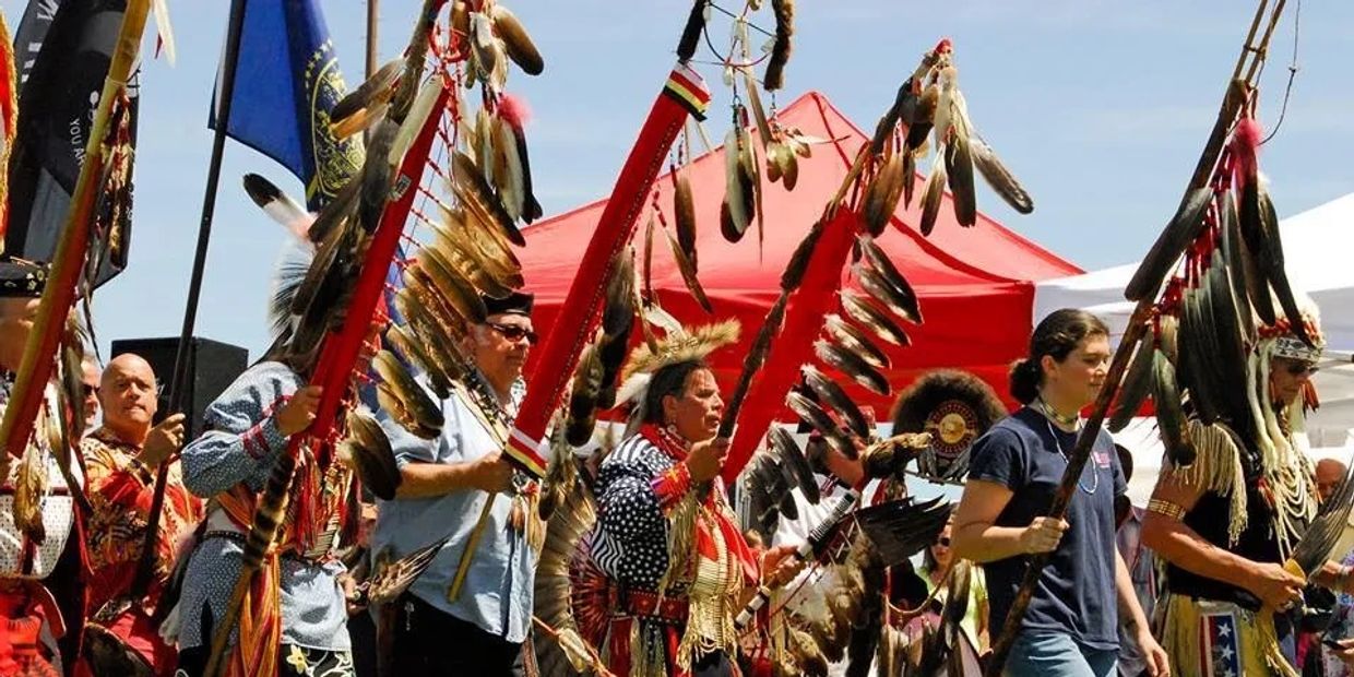 People in traditional Native American attire holding feathered staffs during a cultural event.