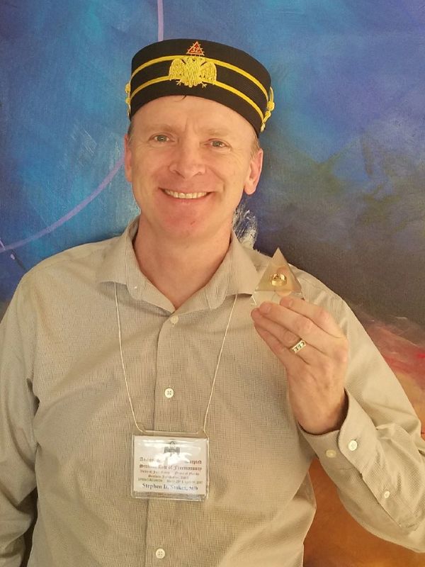 Man in a decorated black hat with a golden emblem, smiling.