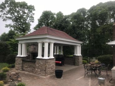 Outdoor stone gazebo with white pillars and a red roof, surrounded by greenery.