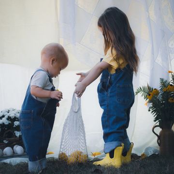 Two children in denim overalls playing with a net bag of lemons outdoors.
