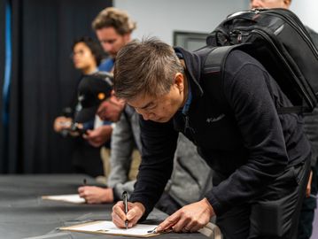 A man intently filling out a form at a table with others in line behind him.