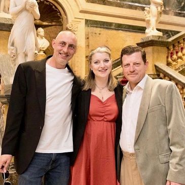 Three people posing together indoors with statues and stairs in the background.