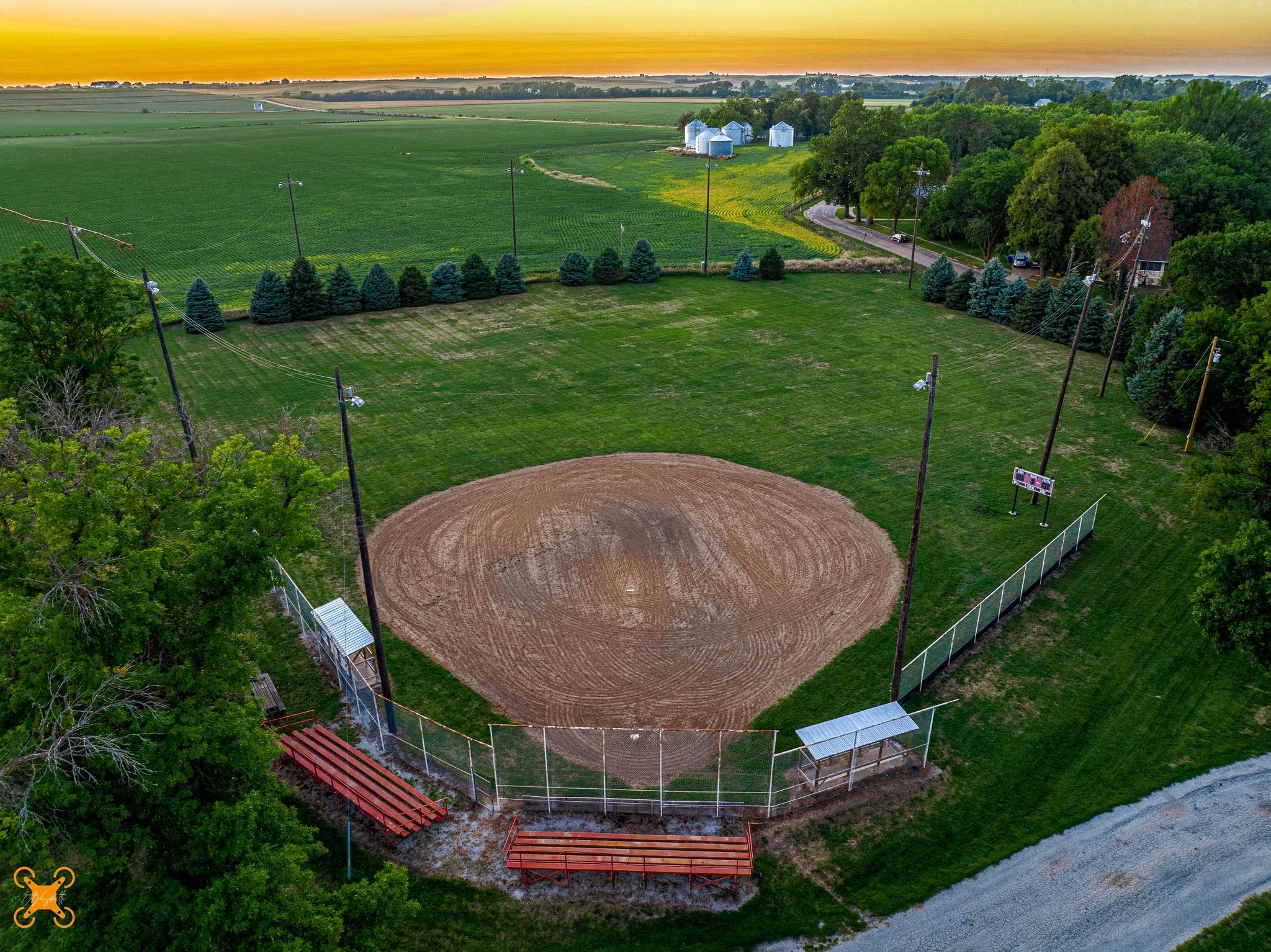 A serene baseball field at sunset surrounded by trees and farmland.